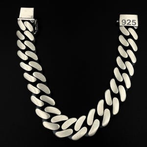 May include: A silver curb chain bracelet with a matte finish. The bracelet features interlocking oval links. The clasp is visible at both ends. The number "925" is stamped on one end, indicating the metal's purity.
