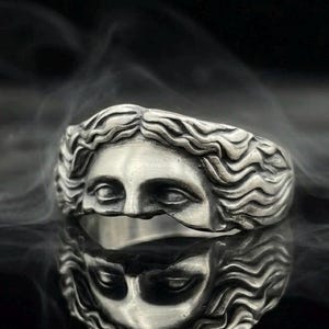May include: A silver ring featuring a sculpted face with detailed, flowing hair. The metal has an oxidized finish that highlights the facial features and the wavy texture of the hair. The ring is shown with a clear reflection on a dark surface.