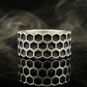 Könnte beinhalten: Ein silberfarbener Ring mit einem sich wiederholenden hexagonalen Wabenmuster auf dem äußeren Band. Die vertieften Bereiche innerhalb jedes Sechsecks haben eine dunkle, texturierte Oberfläche, während die erhabenen Linien glatt sind.