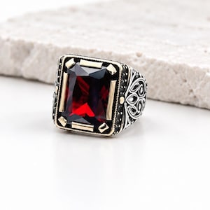 Può includere: Un anello in argento con una grande pietra preziosa rettangolare rosso scuro incastonata in una cornice color oro. La fascia è realizzata in argento ossidato con intricati motivi metallici in stile filigrana sui lati.