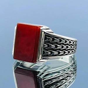 Könnte beinhalten: Ein silberner Ring mit einem rechteckigen, tiefroten Stein. Das Band weist ein detailliertes, sich wiederholendes Muster auf. Der Ring wird auf einer reflektierenden Oberfläche gezeigt, wodurch ein Spiegeleffekt entsteht. Das Design ist kühn und auffällig.