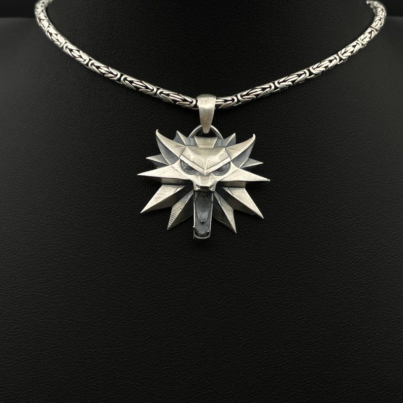 May include: A silver pendant necklace featuring a stylized wolf head emblem. The pendant has a detailed, angular design with a prominent open mouth. The necklace chain is a textured, interwoven pattern. The background is black.