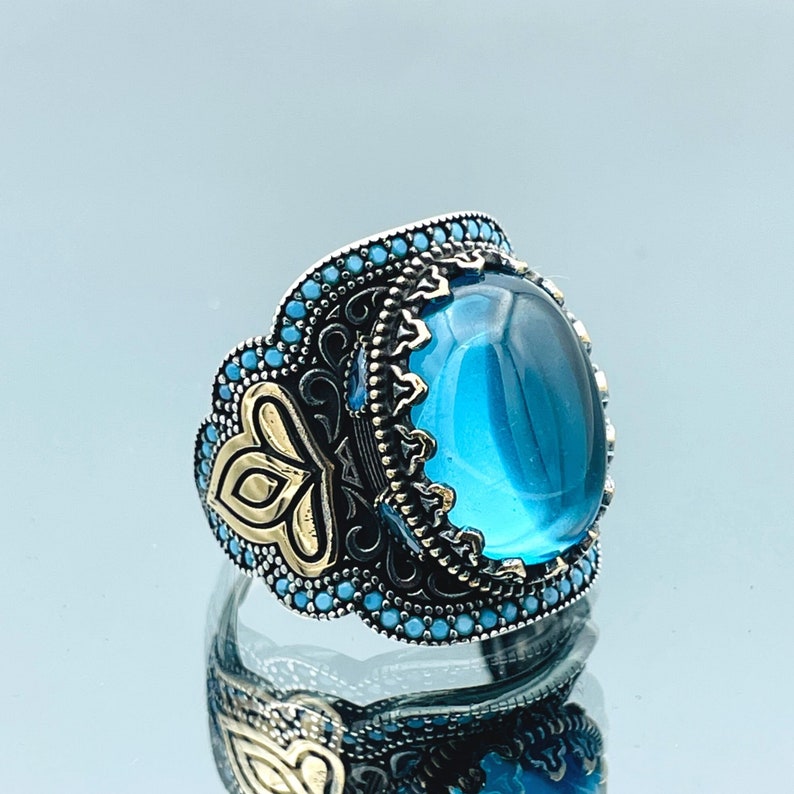 May include: A silver ring with a large blue gemstone set in a detailed silver and gold filigree design. The ring is adorned with small blue stones around the edge.