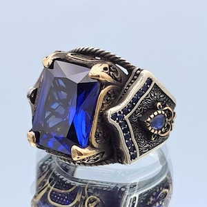 May include: A silver ring with a large, faceted, blue gemstone set in a gold-toned setting. The ring has intricate, detailed designs and smaller blue gemstones around the main stone.