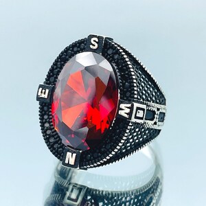 May include: A silver ring with a large red gemstone and black accents. The ring features the letters N, S, E, and W around the gemstone.