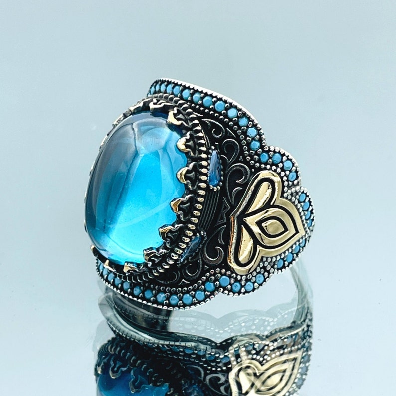 May include: A silver ring with a large, oval-shaped blue gemstone set in a detailed, ornate design. The ring features intricate filigree work and small blue stones around the gemstone.