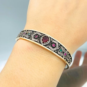 Peut inclure: Un bracelet jonc en argent avec un motif doré aux détails complexes, des pierres précieuses rouges et vertes, et une grande pierre rouge au centre.
