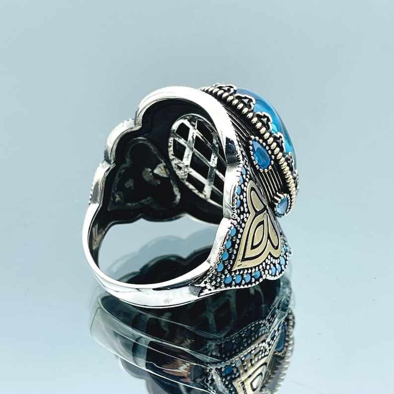 May include: A silver ring with a large blue gemstone surrounded by smaller blue gemstones. The ring has intricate gold detailing and a filigree design.