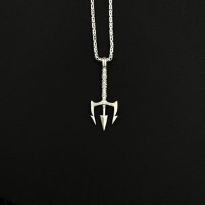 May include: A silver-colored trident pendant necklace. The pendant has a detailed design and is suspended from a matching chain. The necklace is displayed against a black background, highlighting the jewelry's intricate details.