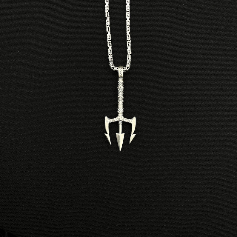 May include: A silver-colored trident pendant necklace. The pendant has a detailed design and is suspended from a matching chain. The necklace is displayed against a black background, highlighting the jewelry's intricate details.
