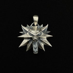 May include: Silver-toned pendant featuring a stylized wolf's head emblem with sharp, angular details. The pendant has a loop at the top for a chain and is set against a dark background. The wolf's head has an open mouth and pointed ears.