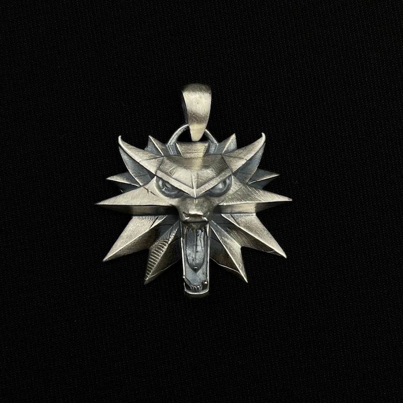 May include: Silver-toned pendant featuring a stylized wolf's head emblem with sharp, angular details. The pendant has a loop at the top for a chain and is set against a dark background. The wolf's head has an open mouth and pointed ears.