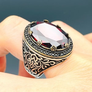 May include: A silver ring featuring a large, oval-shaped, dark red gemstone set in a gold-toned bezel. The ring is adorned with intricate, floral-inspired designs and black accents.