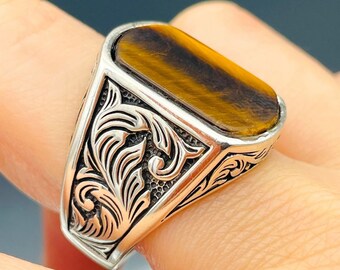 May include: A silver ring with a brown and gold tiger's eye stone set in a rectangular bezel. The ring band is decorated with a detailed floral design.