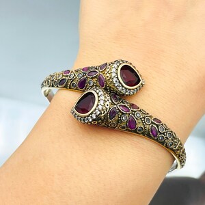 Può includere: Un bracciale dorato ornato con una base argentata, con un design avvolgente. Il bracciale è adornato con pietre preziose color rubino e cristalli trasparenti, creando un contrasto sorprendente. Il design incorpora motivi floreali e geometrici.