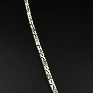 May include: A silver-toned bracelet with a repeating pattern of white, elongated oval links. Each link is accented with a green, diamond-shaped design. The bracelet is displayed against a dark background.