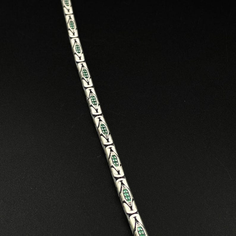 May include: A silver-toned bracelet with a repeating pattern of white, elongated oval links. Each link is accented with a green, diamond-shaped design. The bracelet is displayed against a dark background.