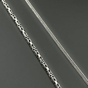 May include: Two silver necklaces on a gray background. One necklace features a Byzantine chain design, while the other has a wheat chain style. Both necklaces are made of a shiny, metallic material.