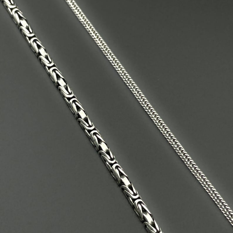 May include: Two silver necklaces on a gray background. One necklace features a Byzantine chain design, while the other has a wheat chain style. Both necklaces are made of a shiny, metallic material.