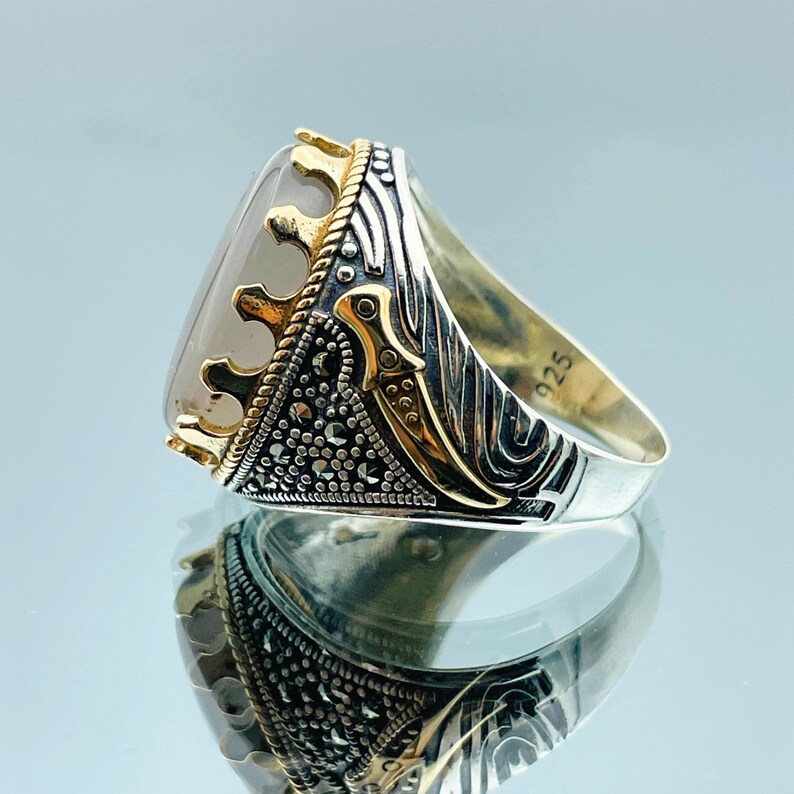 Pode incluir: Um anel de prata com uma pedra grande de forma ovalada, de cor cinza claro, engastada em uma coroa dourada. A banda do anel &eacute; decorada com um design preto e prata e o n&uacute;mero "925" est&aacute; estampado na lateral.