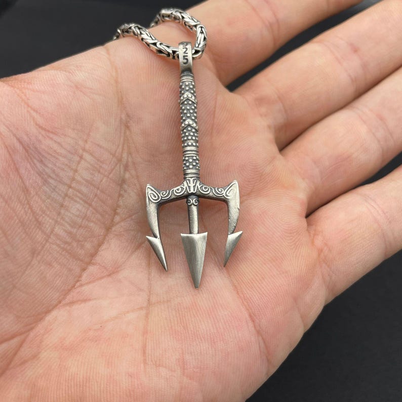 May include: Silver trident pendant necklace. The pendant features a detailed trident design with three prongs and a central spearhead. The necklace has a textured chain and is a stylish accessory.