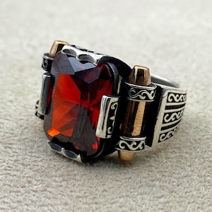 May include: A silver ring with a large, red gemstone set in a square shape. The ring has a decorative design with a gold accent on the side.