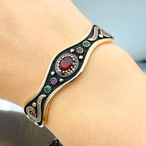 May include: A gold and black bracelet with a red gemstone surrounded by smaller white gemstones. The bracelet has a wave-like design with green and red accents.