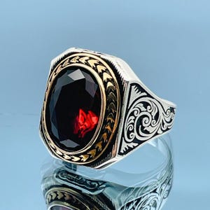 Może przedstawiać: Srebrny pierścionek z owalnym, ciemnoczerwonym kamieniem szlachetnym. Kamień jest osadzony w złotej oprawie. Pierścionek ma ozdobne, grawerowane detale po bokach i złoty akcent wokół kamienia.