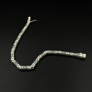 May include: A silver bracelet with a series of oval links, each containing a row of small, emerald-green stones. The bracelet is laid out on a black surface, with the clasp visible at the end. The clasp is marked with "925".