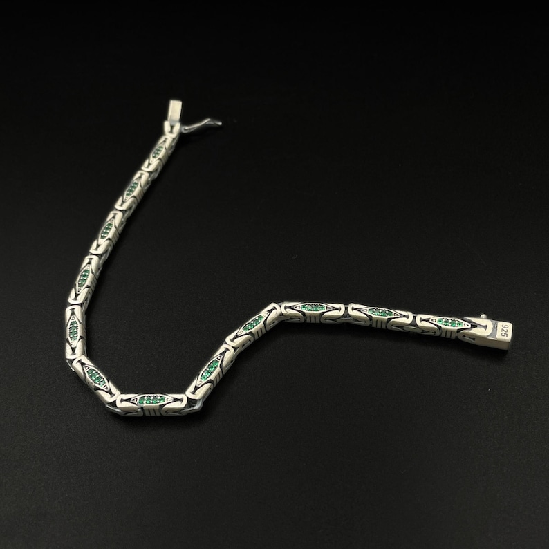 May include: A silver bracelet with a series of oval links, each containing a row of small, emerald-green stones. The bracelet is laid out on a black surface, with the clasp visible at the end. The clasp is marked with "925".