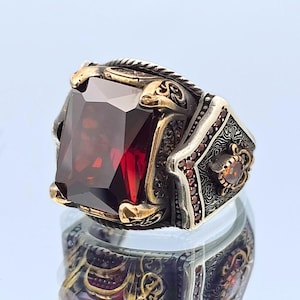 Peut inclure: Une bague en argent avec une grande pierre précieuse rouge sertie dans un sertissage doré. La bague est décorée de motifs complexes et détaillés.