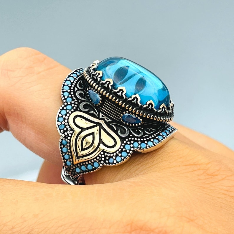 May include: A silver ring with a large blue gemstone surrounded by smaller blue gemstones and intricate gold detailing.
