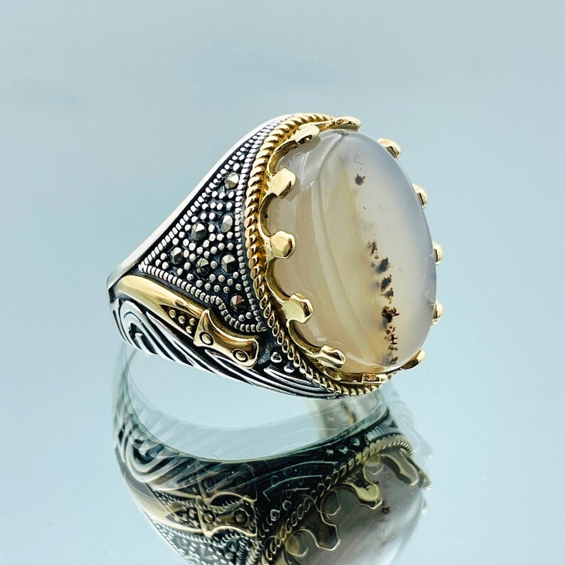 Pode incluir: Um anel de prata com um design de coroa dourada e uma pedra de &aacute;gata branca. O anel tem um design detalhado com detalhes pretos.