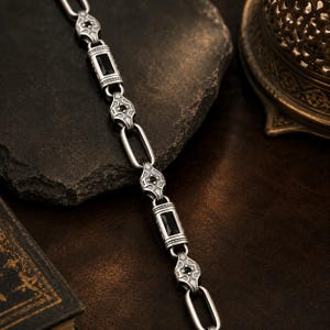 Könnte beinhalten: Ein silbernes Kettenarmband mit abwechselnden rechteckigen Gliedern mit schwarzen Steinen und verzierten, schildförmigen Gliedern, die mit kleinen schwarzen Steinen besetzt sind. Das Metall hat ein mattes, oxidiertes Finish mit Gravuren.