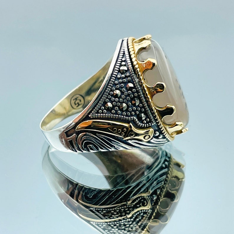 Pode incluir: Um anel de prata e ouro com uma pedra cinza engastada em uma moldura em forma de coroa. O anel &eacute; decorado com pedras pretas de marcasita.