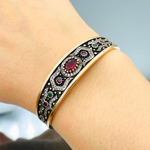 May include: A gold-toned bangle bracelet with an intricate design featuring red, green, and white gemstones. The bracelet has a black oxidised finish and a detailed floral pattern.