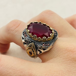 May include: A silver and gold ring with a large, red gemstone. The ring has an intricate design with small red gemstones around the main stone.
