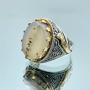Pode incluir: Um anel de prata com uma grande pedra de &aacute;gata ovalada engastada em uma moldura dourada em forma de coroa. A banda &eacute; decorada com um design preto e prateado e uma pequena adaga dourada.
