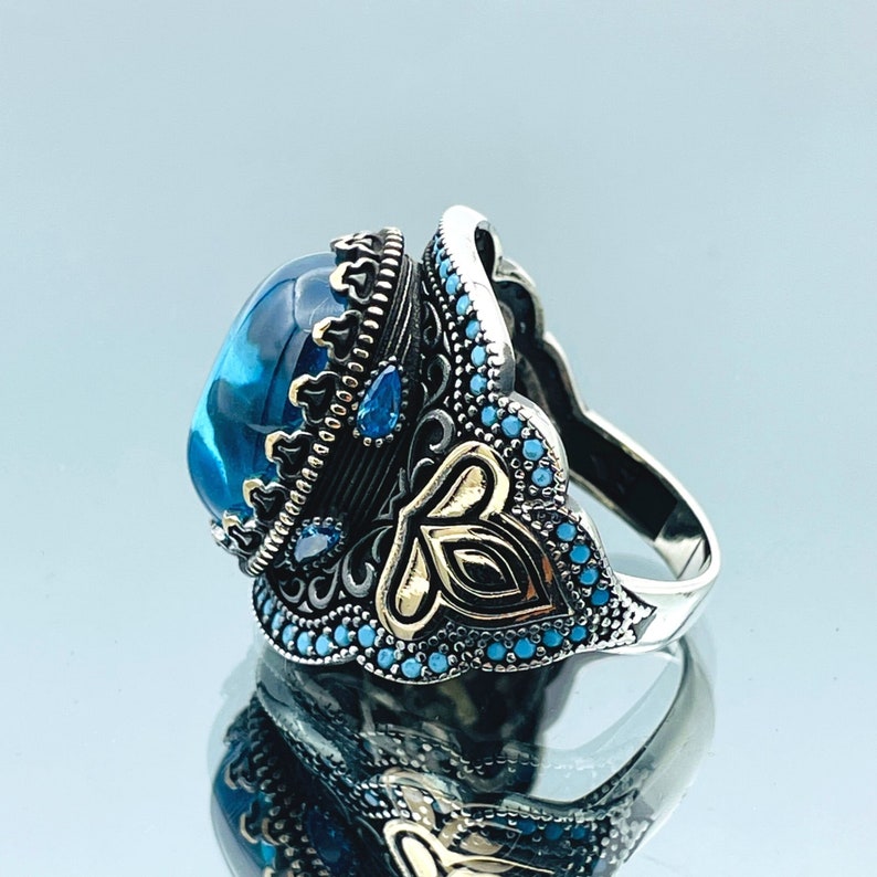 May include: A silver ring with a large blue gemstone surrounded by smaller blue gemstones and gold accents. The ring has a detailed design with intricate carvings.