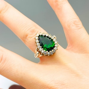 Può includere: Un anello d'oro con una grande pietra preziosa verde a forma di pera circondata da piccole pietre preziose chiare.