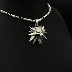 May include: A silver pendant necklace featuring a stylized wolf head emblem. The pendant has a detailed, geometric design with sharp angles and a textured finish. The chain is a detailed, woven pattern.