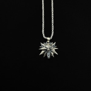 May include: Silver-colored pendant necklace featuring a wolf head emblem. The pendant has a detailed, angular design with a central wolf face and surrounding stylized rays. The chain is a linked design, and the background is black.