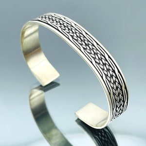 Könnte beinhalten: Ein silbernes Armband mit einem geflochtenen Design. Das Armband ist offen und hat eine glatte, polierte Oberfläche.