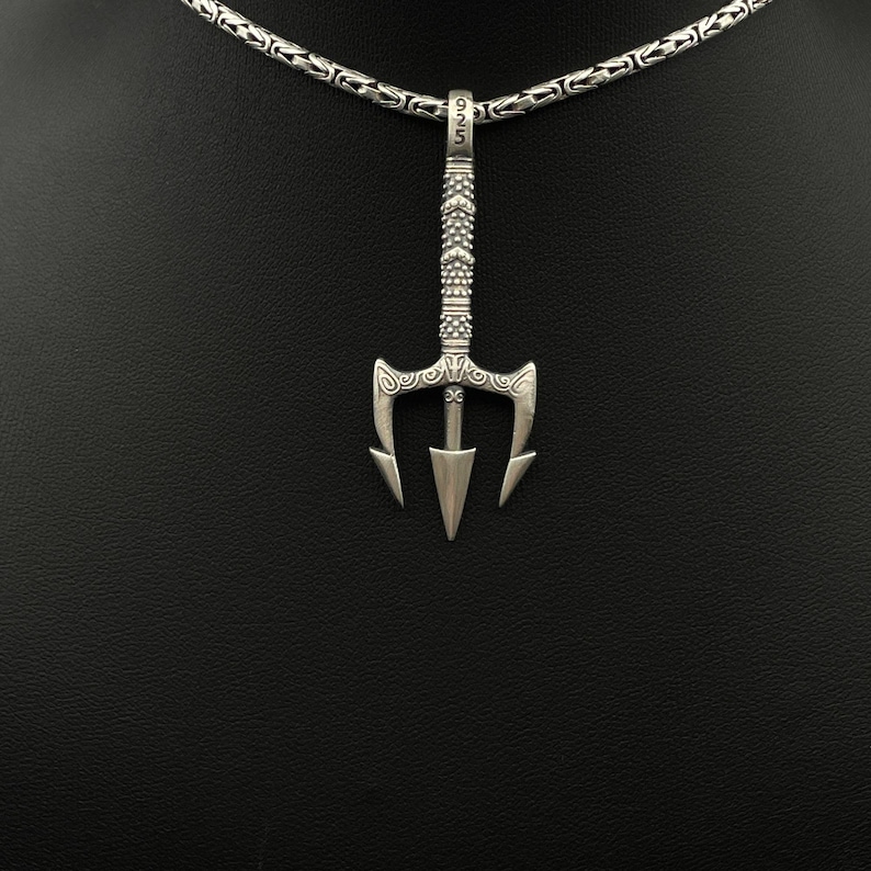 May include: A silver trident pendant necklace. The pendant is a detailed trident design with a textured handle and a pointed tip. The necklace chain is a linked design. The pendant is suspended from the chain.
