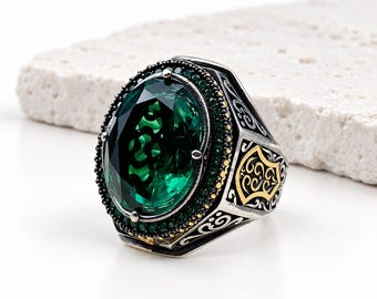 May include: A silver ring featuring a large oval-cut emerald green stone surrounded by a border of small green crystals. The band is crafted from oxidized silver with intricate scrollwork engravings and gold-toned accents on the sides.