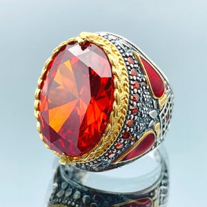 May include: A silver ring with a large, red, faceted gemstone set in a gold-toned bezel. The ring is decorated with intricate silver and red enamel details.