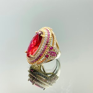 May include: A gold ring with a large oval-shaped red gemstone surrounded by smaller clear and pink gemstones.