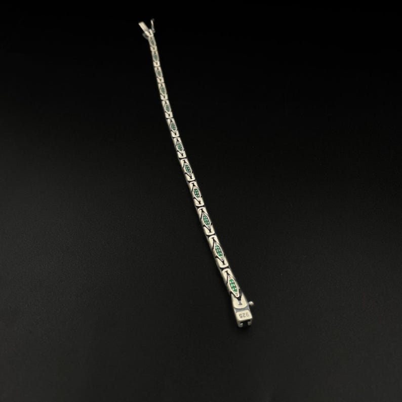 May include: A silver bracelet with a series of elongated, leaf-shaped links, each containing a green stone. The bracelet is laid out diagonally against a black background. The clasp is visible at the bottom, with a 925 stamp.