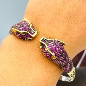 Peut inclure: Un bracelet en argent avec deux têtes de panthère à chaque extrémité. Les têtes de panthère sont recouvertes de petites pierres précieuses violettes. Le bracelet est ouvert et présente un design en chaîne sur le dos.