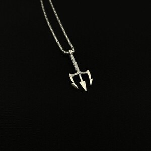 May include: A silver-colored pendant necklace featuring a trident design. The trident has three sharp prongs and is suspended from a delicate chain. The necklace is displayed against a black background, highlighting the intricate details of the jewelry.